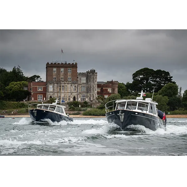 Two boats sail on a body of water with a large historic building and trees in the background under a cloudy sky, where yacht insurance could provide peace of mind for such serene journeys.