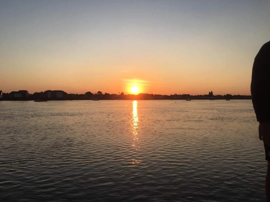 A sunset over a calm body of water with a silhouetted shoreline in the distance and a partial view of a person on the right.