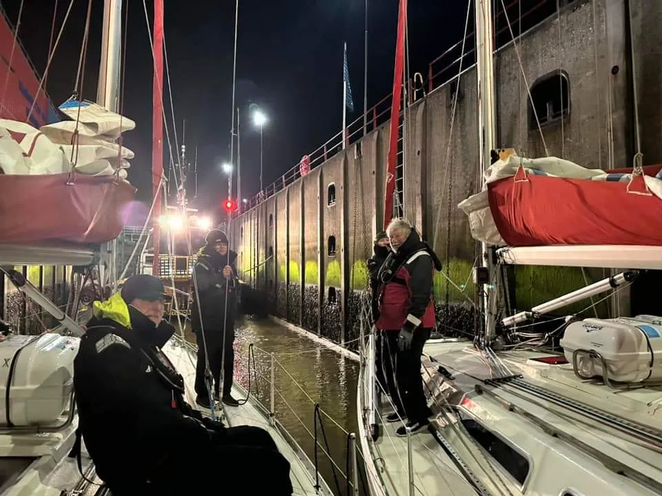 People standing on two sailboats in a lock at night, holding ropes and wearing jackets, with high concrete walls surrounding them.