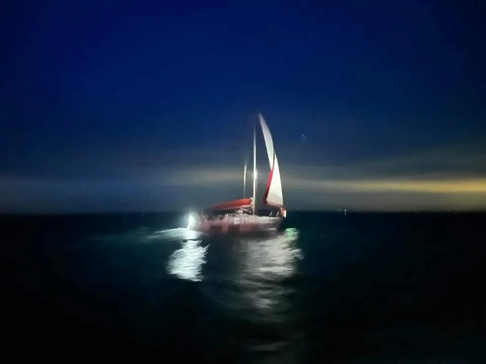 A sailboat with red and white sails moves through dark waters at dusk, illuminated by a light.