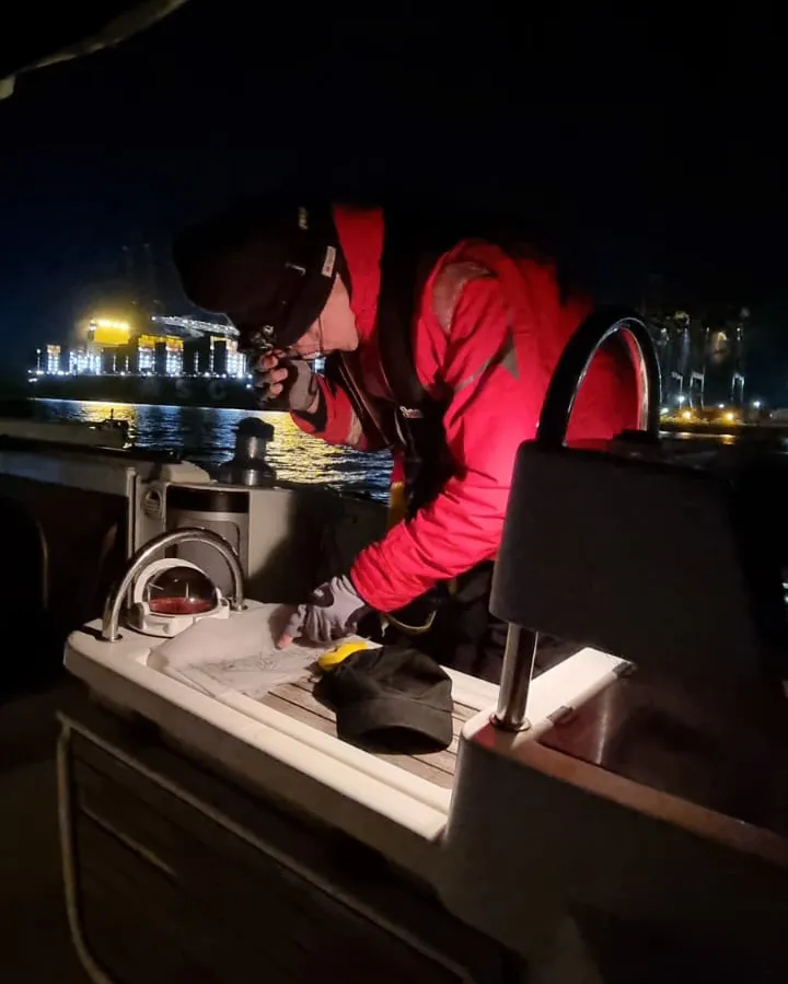 Person in red jacket and black hat working on a boat at night, illuminated by a light, with a cityscape in the background.