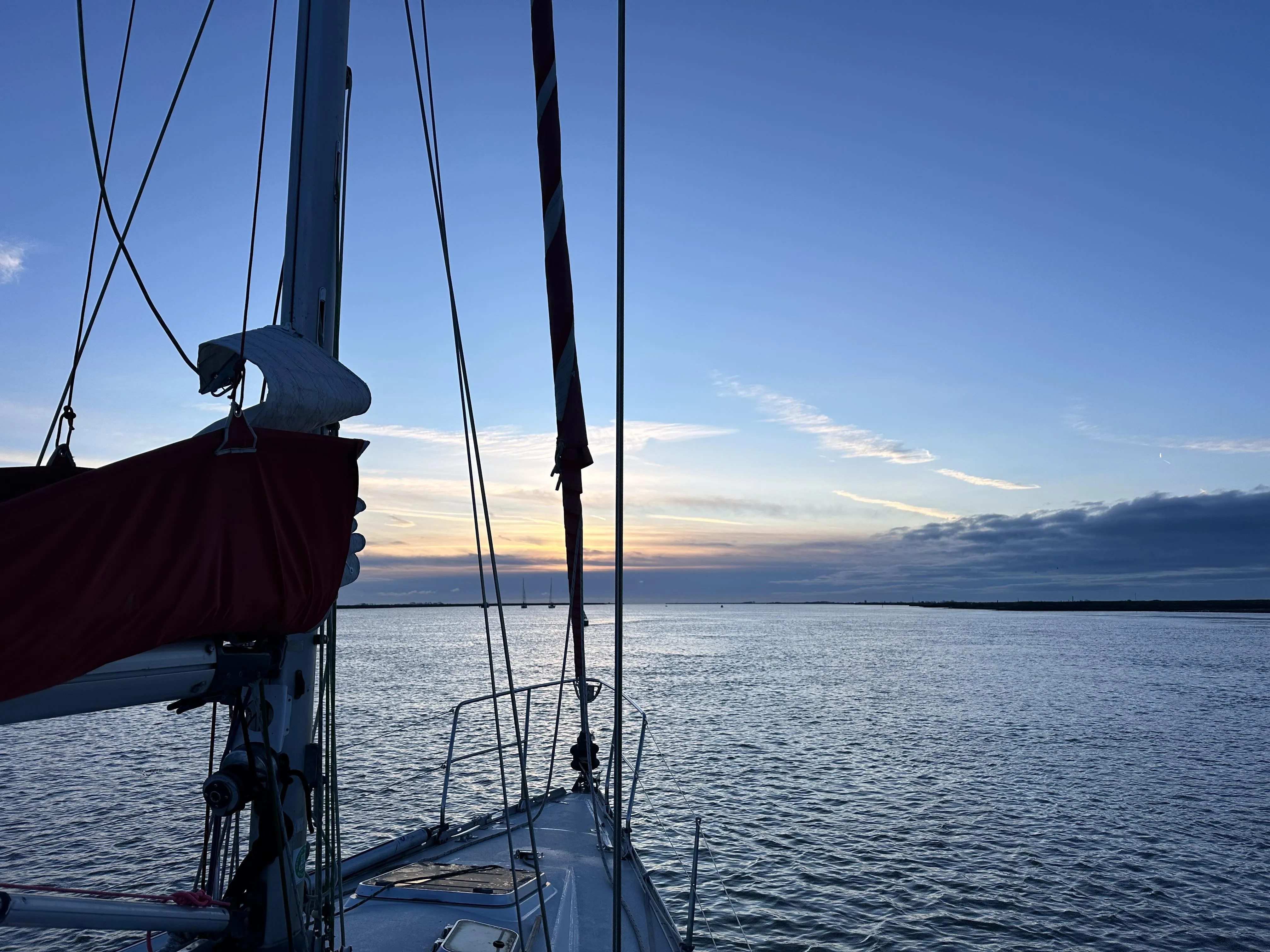 A sailboat with a red sail is on calm waters during sunset, with a horizon of dark clouds and a clear sky above.