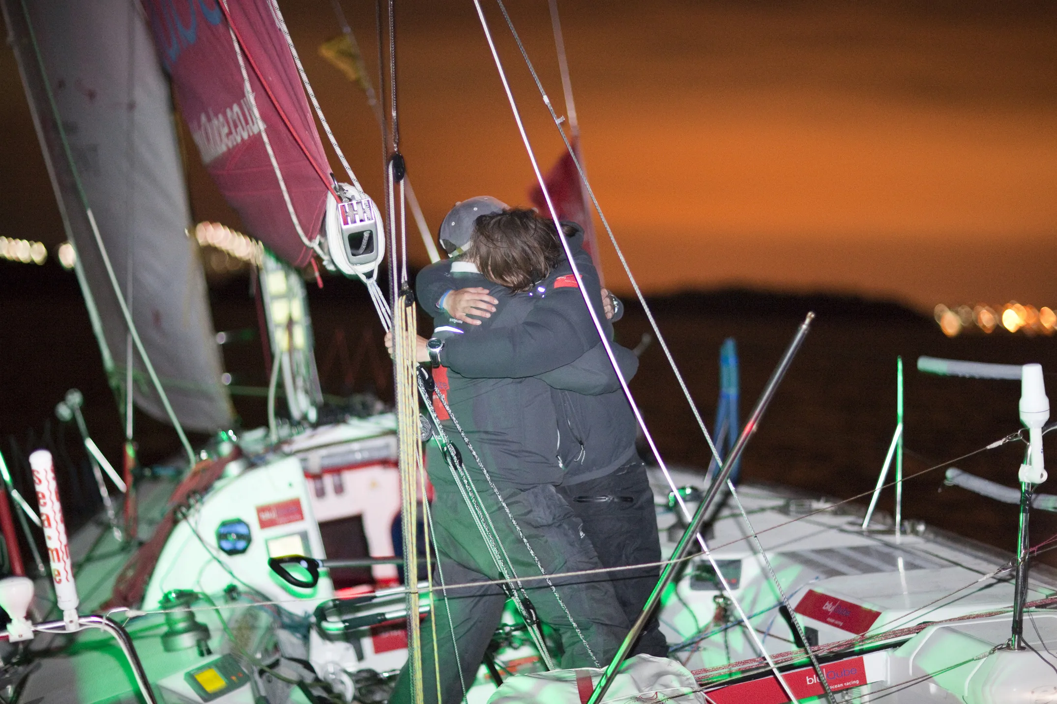 Two individuals share an embrace on a sailboat at night, under a sky lit by an orange glow.