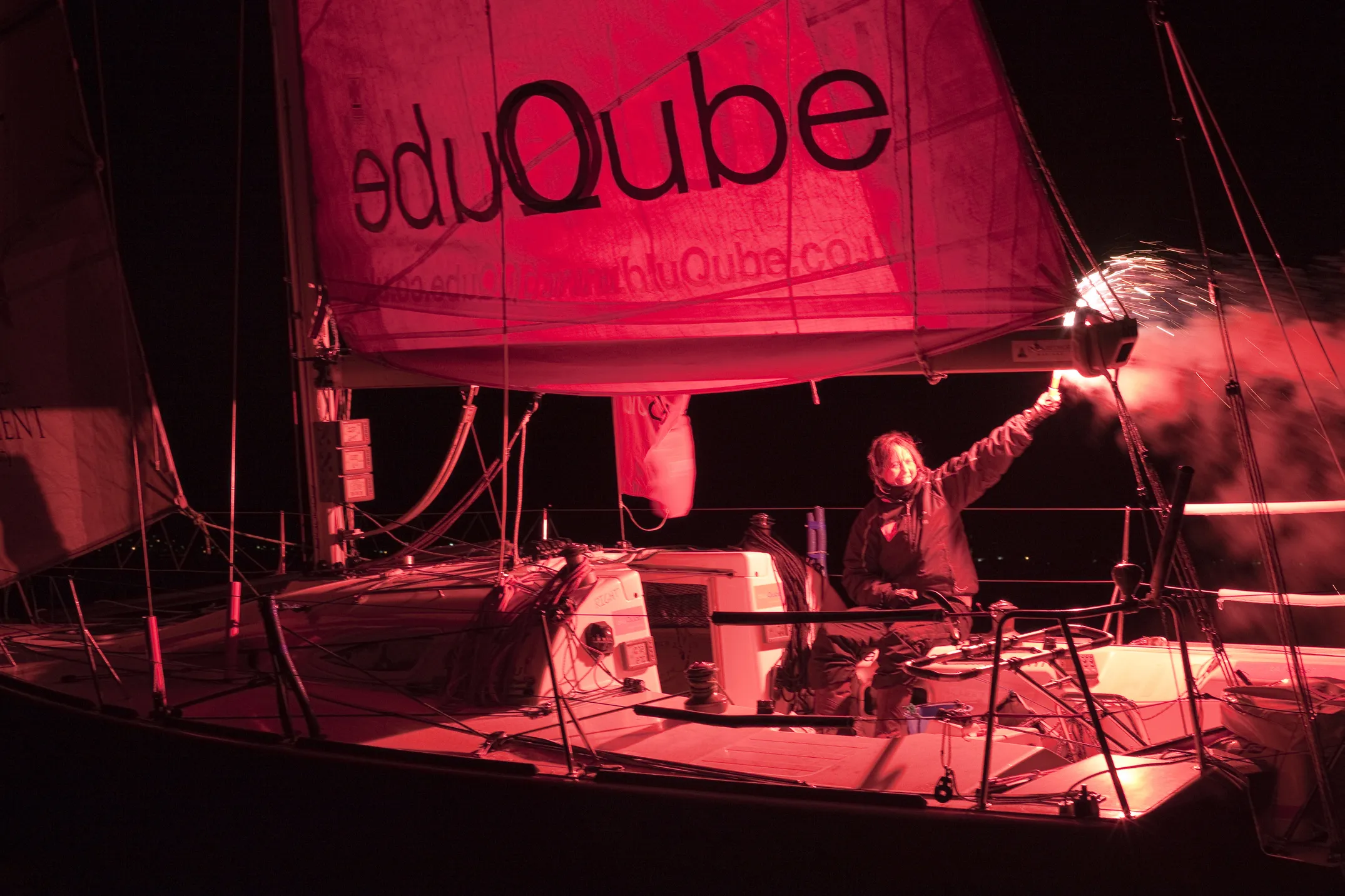 A person on a sailboat at night holding a flare. The sail reads 