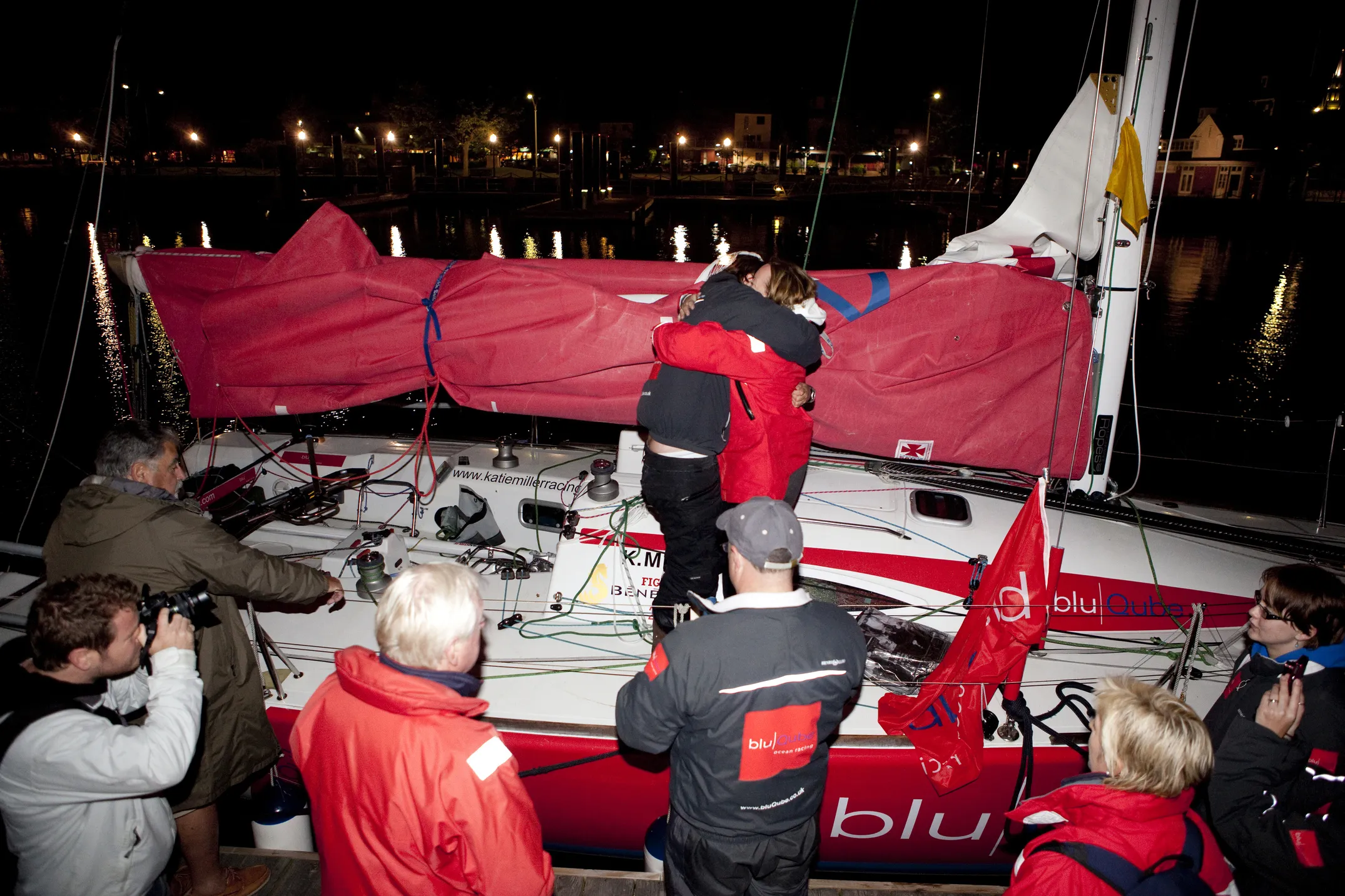 People in sailing gear embrace on a docked sailboat at night, with others nearby capturing the moment and observing.