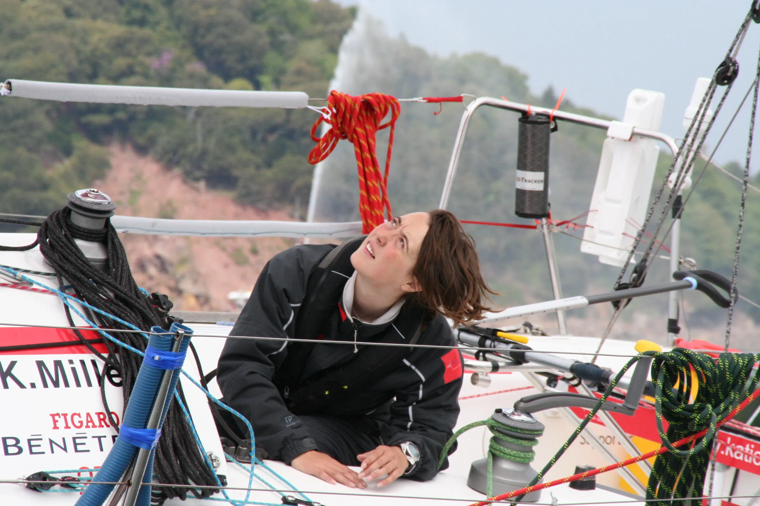 A person in a sailing suit aboard a boat, looking upwards, surrounded by ropes and sailing equipment, with trees in the background.