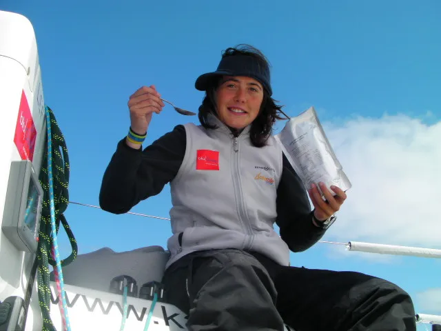 Person in outdoor gear eating from a packet while sitting on a boat deck, with ropes and equipment visible in the background.