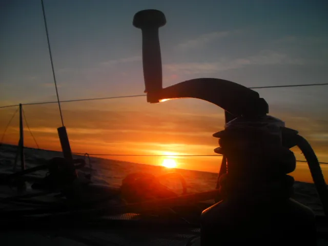 Sunset viewed from a sailing boat, with a winch and ropes in the foreground against the colorful sky and horizon.