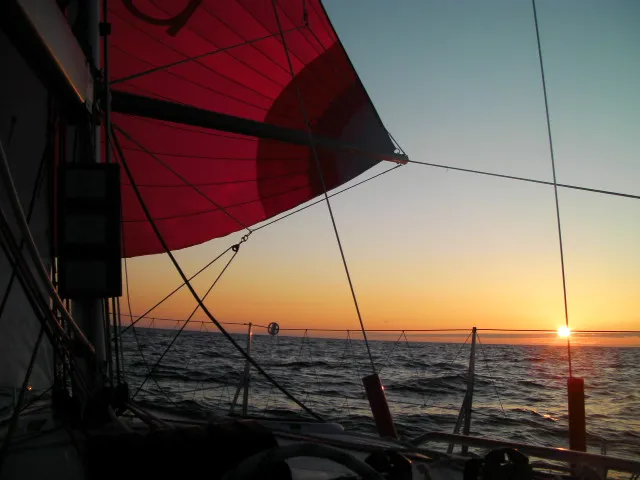 Sailboat on the ocean with a red sail at sunset. The horizon and the sun are visible in the background.