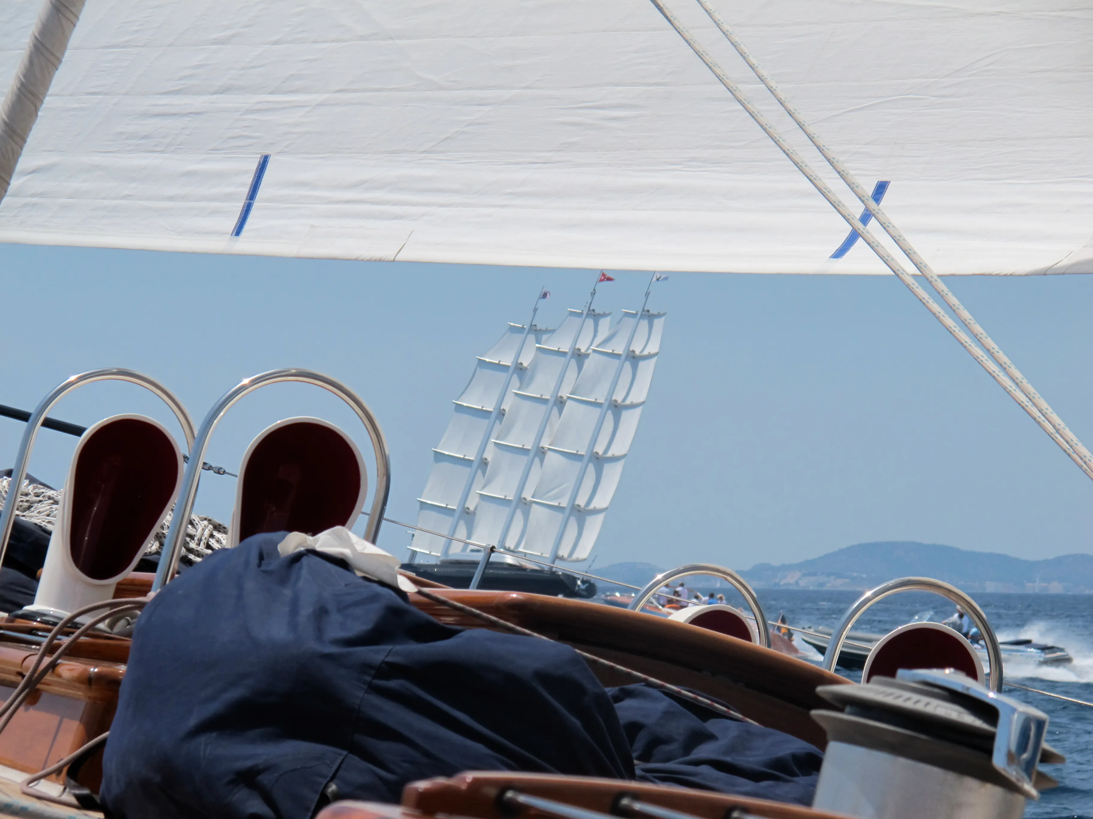 View from a sailboat in the foreground with a larger sailboat, possibly under the command of a superyacht captain, in the distance, all set against a vibrant blue sky.