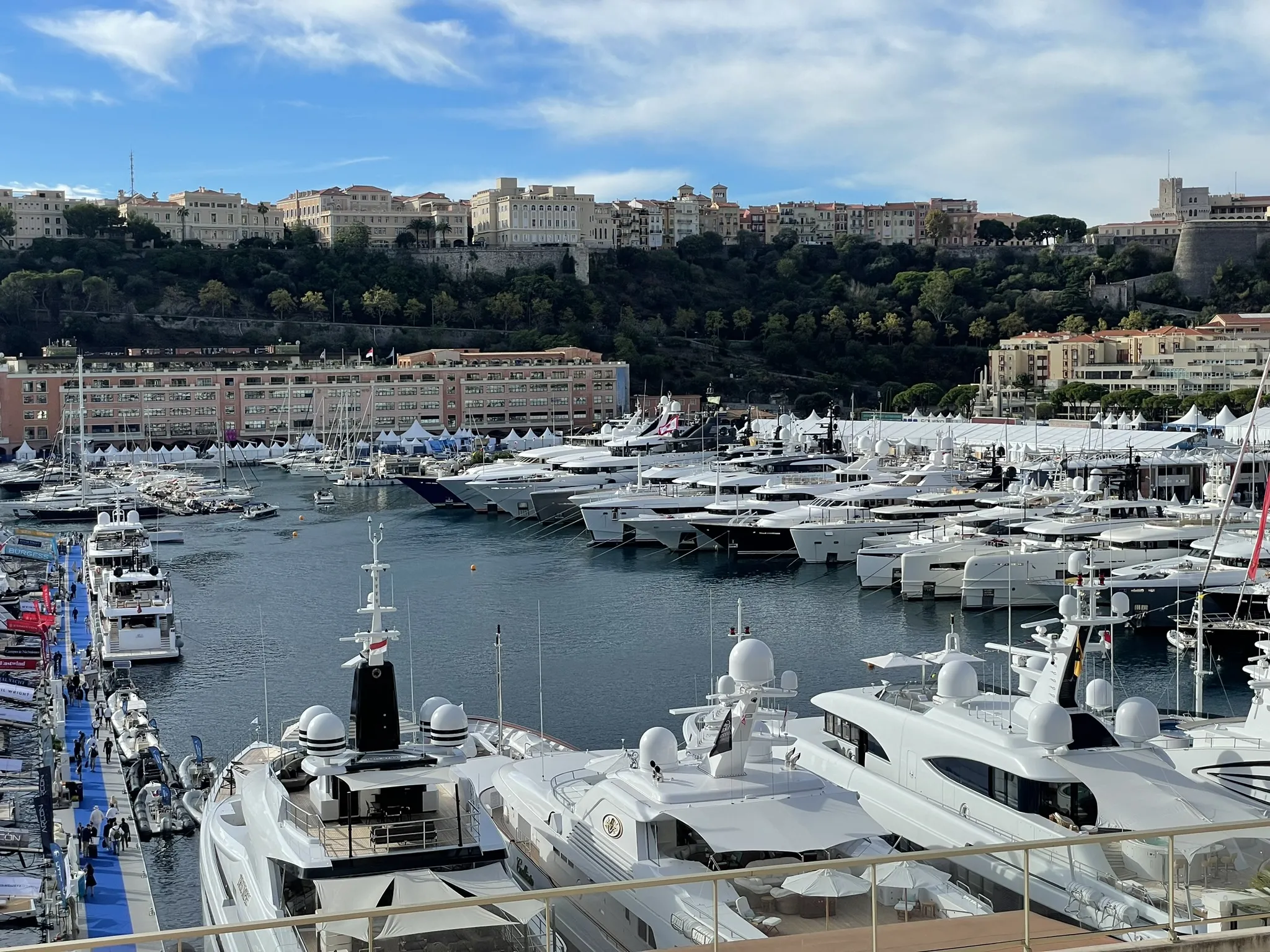 A marina filled with numerous luxury yachts docked in the water, overseen by skilled superyacht captains, with a hillside cityscape and dense greenery in the background.