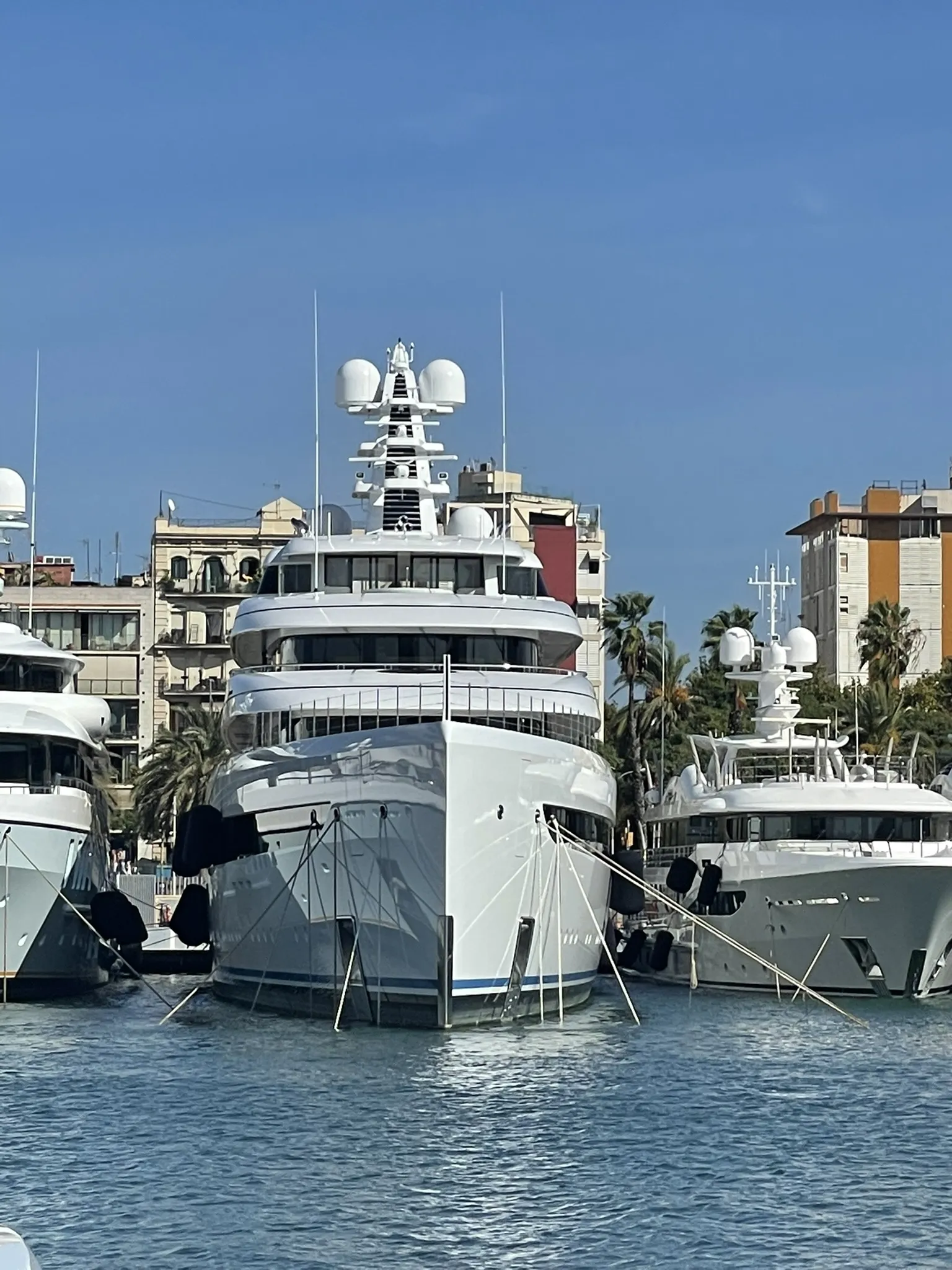 Three large luxury yachts, each under the watchful eye of a seasoned superyacht captain, docked side by side in a marina. Buildings and palm trees grace the background under a clear blue sky, setting a scene of serene opulence.