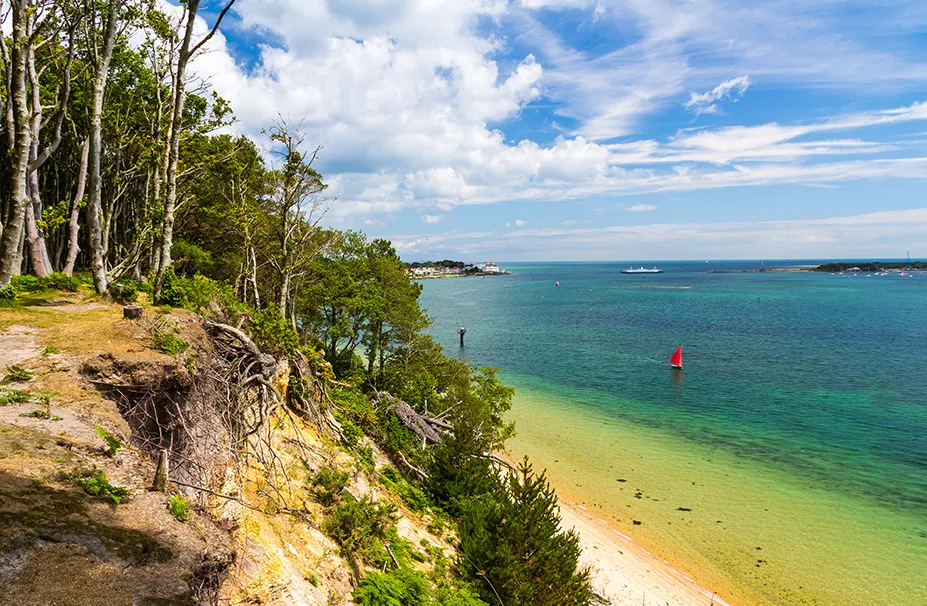 A coastal scene with a forested cliff, clear blue water, a sandy beach, and a small red sailboat in the distance under a partly cloudy sky, reminiscent of the tranquil moments enjoyed by a superyacht captain.