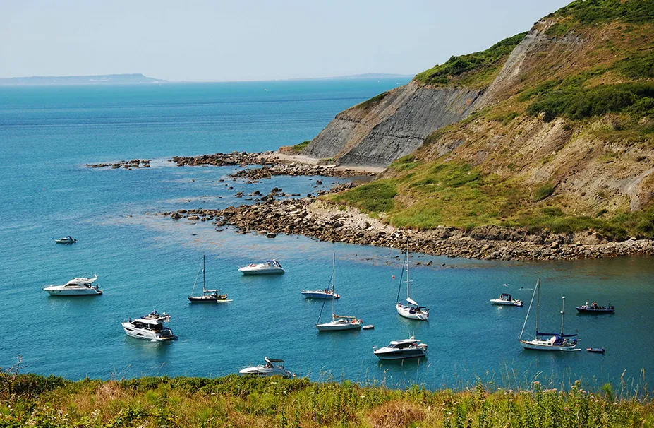 Several boats and yachts, guided by seasoned superyacht captains, anchor in a calm bay near a rocky coastline with lush green hills and clear blue water.