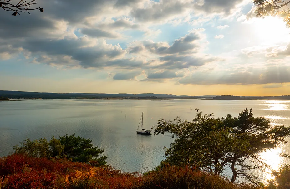 A sailboat navigated by a superyacht captain glides on a calm lake, with sunlight breaking through the partially cloudy sky and casting reflections on the water, surrounded by trees and distant hills.