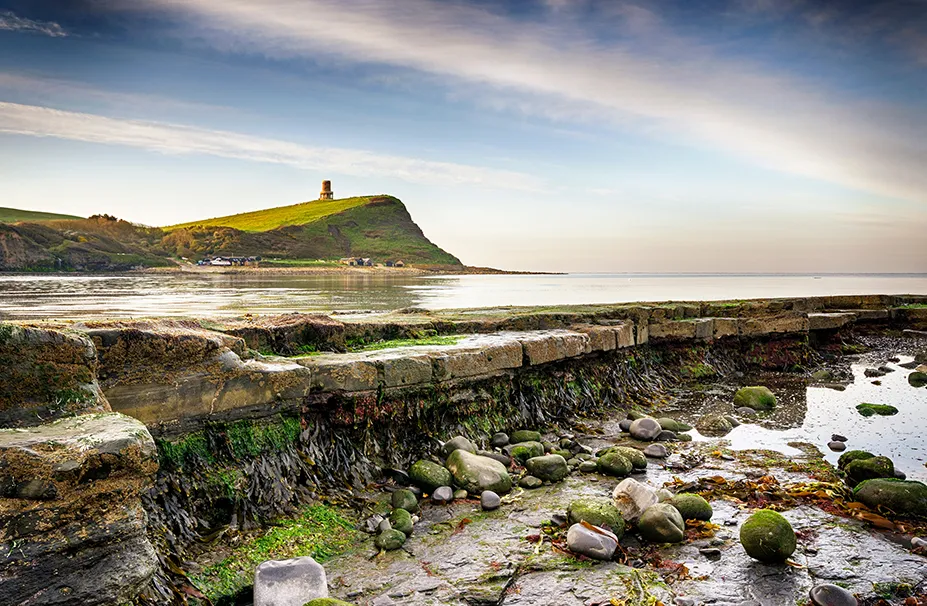 Coastal landscape with a stone pier in the foreground, rocky shoreline, and a hill with a small tower under a partially cloudy sky, as if awaiting the keen eye of a superyacht captain navigating the serene waters.