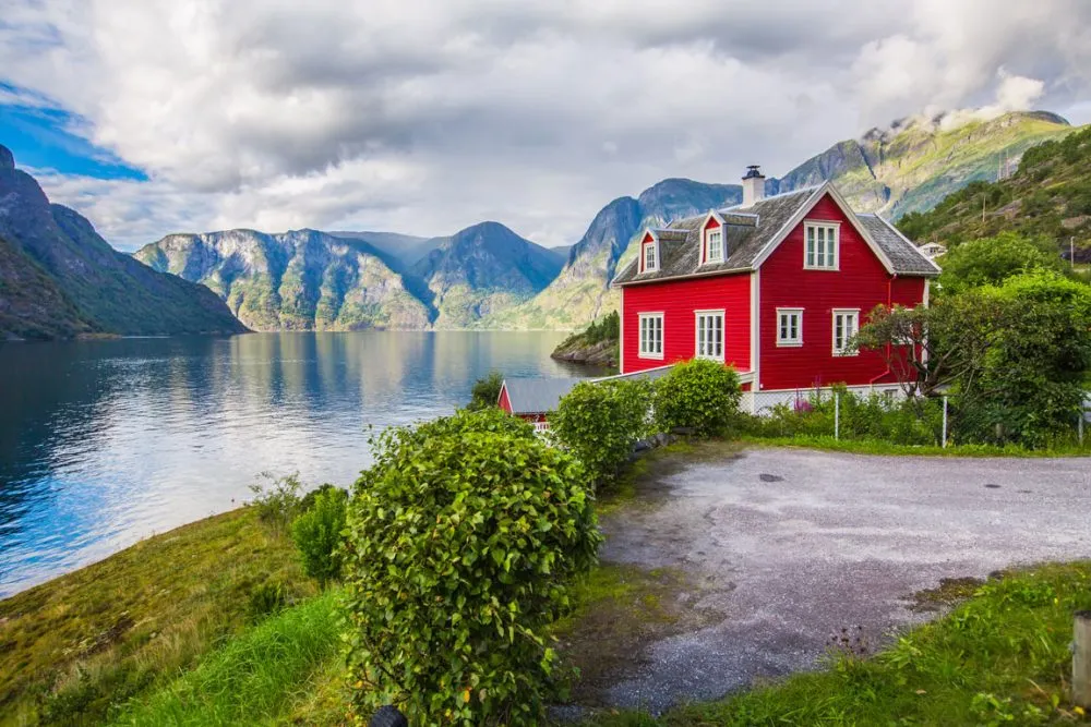 A red house by a calm fjord, renowned among sailing destinations, surrounded by green foliage with majestic mountains in the background under a cloudy sky.