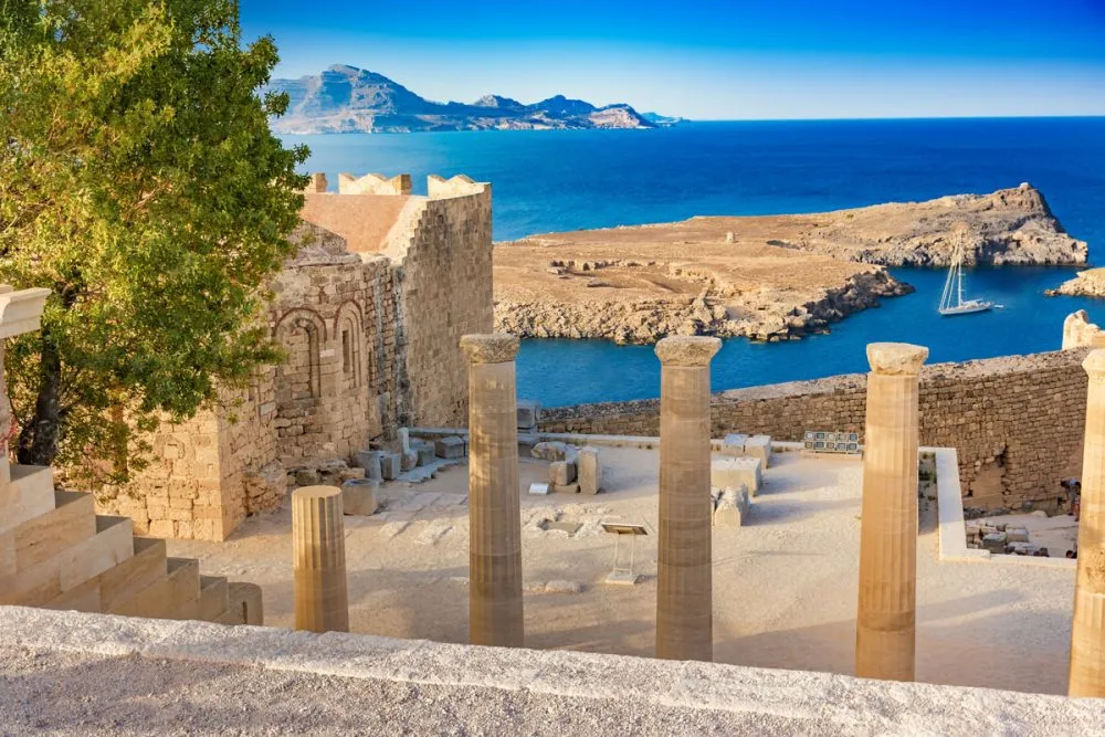 Ancient stone columns and ruins overlook a rocky coastline and blue sea, under a clear sky, making it one of the most scenic sailing destinations.