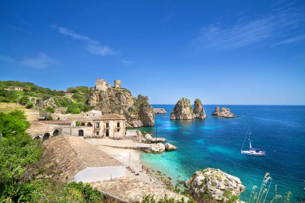 A coastal scene reveals a stone building beside turquoise seas and striking rock formations, with a sailboat anchored nearby under a clear blue sky, epitomizing one of the finest sailing destinations.